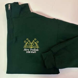 K&ouml;nnte beinhalten: Ein dunkelgr&uuml;ner Pullover mit einem Rei&szlig;verschluss an der Vorderseite. Der Pullover hat ein gesticktes Design eines Buches mit Sternen dar&uuml;ber. Der Text "Mrs. Pollack IHM Staff" ist unter dem Buch gestickt.