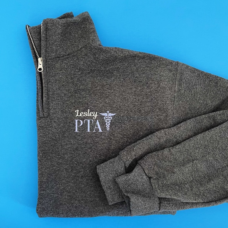 May include: A dark gray quarter-zip sweatshirt with the embroidered text "Lesley PTA" and a medical symbol in white. The sweatshirt has a silver zipper and long sleeves. The background is a solid blue.