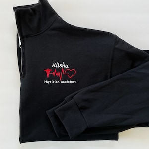 Peut inclure: Sweat-shirt noir &agrave; fermeture &eacute;clair quart de zip avec texte et graphiques brod&eacute;s. Le motif comprend le nom "Alisha", un symbole m&eacute;dical, une ligne de battement de c&oelig;ur et un c&oelig;ur. Les mots "Physician Assistant" sont &eacute;galement brod&eacute;s en blanc.