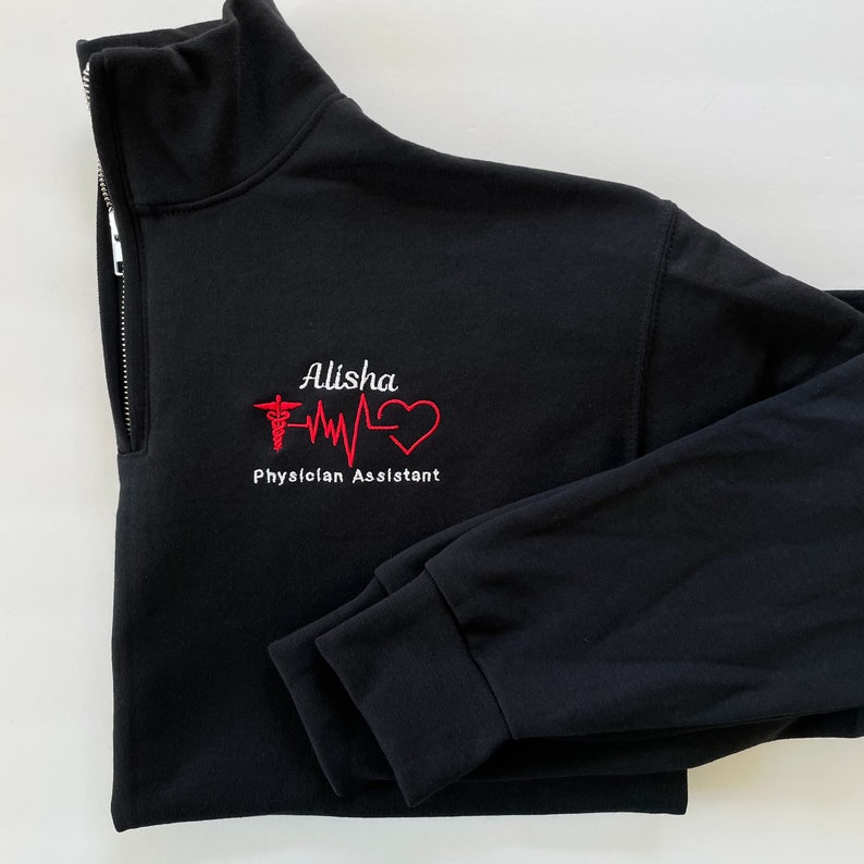 Peut inclure: Sweat-shirt noir &agrave; fermeture &eacute;clair quart de zip avec texte et graphiques brod&eacute;s. Le motif comprend le nom "Alisha", un symbole m&eacute;dical, une ligne de battement de c&oelig;ur et un c&oelig;ur. Les mots "Physician Assistant" sont &eacute;galement brod&eacute;s en blanc.