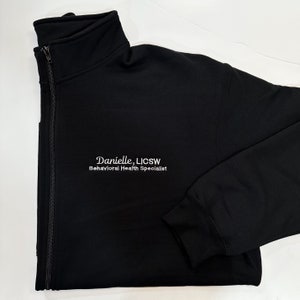 May include: Black zip-up jacket with a high collar. The jacket features the embroidered text "Danielle, LICSW Behavioral Health Specialist" in white. The jacket is made of a soft material and has a full-length zipper.