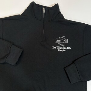 Pode incluir: Camisola preta com fecho de correr de um quarto de gola com texto e gr&aacute;ficos brancos. O texto diz "Dr. Wilson, MD Allergist" abaixo de um gr&aacute;fico de um punho, seringa e outras ferramentas m&eacute;dicas. A camisola tem gola alta e punhos canelados.