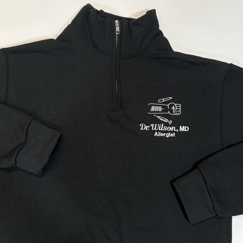 Pode incluir: Camisola preta com fecho de correr de um quarto de gola com texto e gr&aacute;ficos brancos. O texto diz "Dr. Wilson, MD Allergist" abaixo de um gr&aacute;fico de um punho, seringa e outras ferramentas m&eacute;dicas. A camisola tem gola alta e punhos canelados.