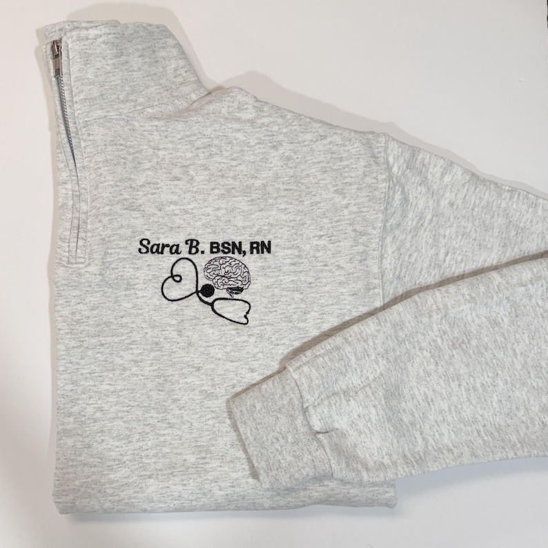 May include: A light gray quarter-zip sweatshirt with the embroidered text "Sara B. BSN, RN" and a stethoscope design with a brain graphic. The sweatshirt has a high collar and a silver zipper.