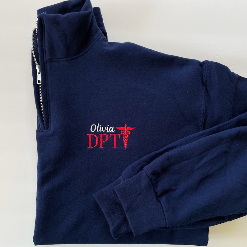 Puede incluir: Sudadera azul marino con cremallera de un cuarto de cuello, con el texto bordado "Olivia DPT" en blanco y rojo, junto con un s&iacute;mbolo de caduceo rojo. La sudadera tiene cuello alto y mangas largas.