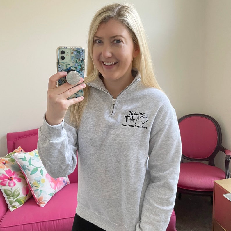 Peut inclure: Un sweat-shirt gris clair &agrave; fermeture &eacute;clair quart de col avec le nom "Kristina" et les mots "Physician Assistant" imprim&eacute;s en noir. Le motif comprend un symbole m&eacute;dical et un graphique de battement de c&oelig;ur. Le sweat-shirt a un col haut.