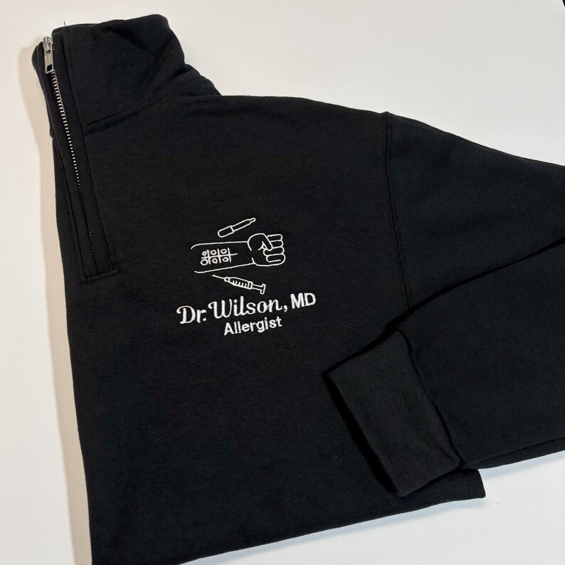 Pode incluir: Camisola preta com fecho de correr de um quarto de gola e gr&aacute;fico branco de um punho, seringa e frascos. O texto "Dr. Wilson, MD Allergist" est&aacute; impresso abaixo. A camisola tem gola alta e punhos canelados.