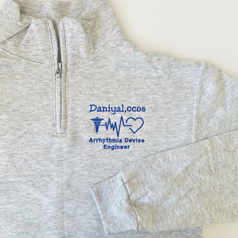 Peut inclure: Un sweat-shirt gris clair &agrave; fermeture &eacute;clair quart de zip avec un motif brod&eacute; bleu. Le motif comprend le nom "Daniyal, ccds", un symbole m&eacute;dical, une ligne de battement de c&oelig;ur, un c&oelig;ur et les mots "Arrhythmia Device Engineer".