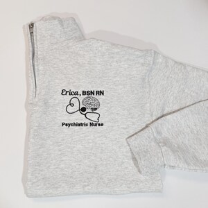 May include: A light gray quarter-zip sweatshirt with the text "Erica, BSN RN" and "Psychiatric Nurse" printed in black. A stethoscope and brain graphic is also printed on the front. The sweatshirt has a high collar and a silver zipper.