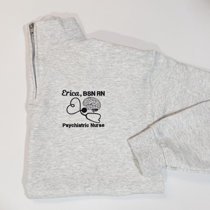 May include: A light gray quarter-zip sweatshirt with the text "Erica, BSN RN" and "Psychiatric Nurse" printed in black. A stethoscope and brain graphic is also printed on the front. The sweatshirt has a high collar and a silver zipper.