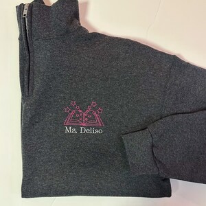 K&ouml;nnte beinhalten: Ein dunkelgrauer Pullover mit einem rosa gestickten Design eines Buches mit Sternen dar&uuml;ber. Der Text "Ms. Deliso" ist unter dem Buch gestickt.