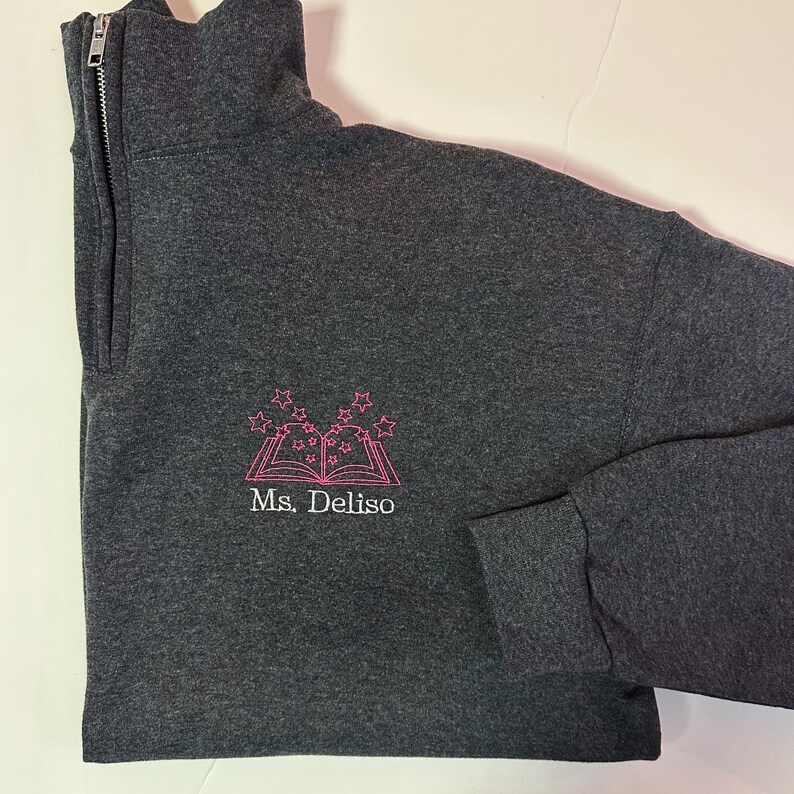 K&ouml;nnte beinhalten: Ein dunkelgrauer Pullover mit einem rosa gestickten Design eines Buches mit Sternen dar&uuml;ber. Der Text "Ms. Deliso" ist unter dem Buch gestickt.