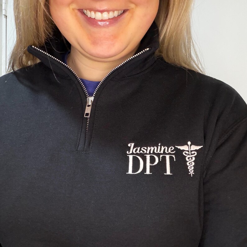 Puede incluir: Sudadera negra con cremallera de cuarto de cremallera con el texto "Jasmine DPT" en escritura blanca y un s&iacute;mbolo m&eacute;dico. La sudadera tiene una cremallera y un cuello acanalado.