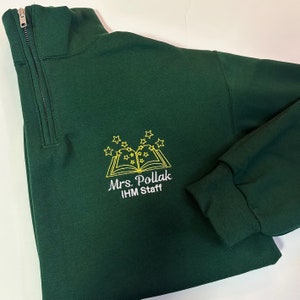 K&ouml;nnte beinhalten: Ein dunkelgr&uuml;ner Pullover mit einem Viertelrei&szlig;verschluss. Der Pullover hat ein gesticktes Design eines Buches mit Sternen dar&uuml;ber und dem Text "Mrs. Pollack IHM Staff".
