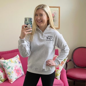 Peut inclure: Un sweat-shirt gris clair avec un col montant zipp&eacute;. Le sweat-shirt porte le nom "Kristina" au-dessus d'un symbole m&eacute;dical et les mots "Physician Assistant" en noir. La personne porte des leggings noirs.