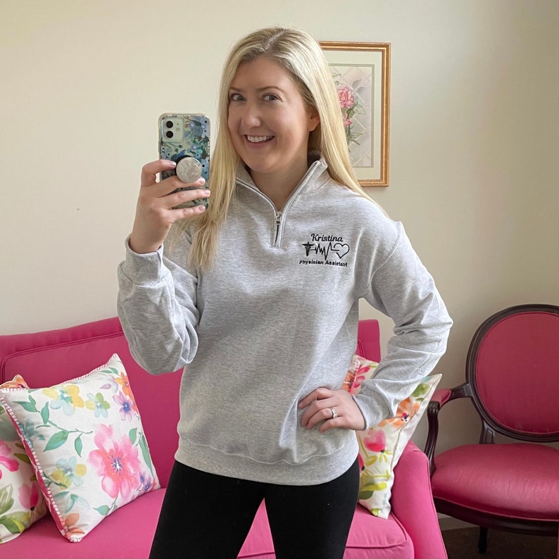 Peut inclure: Un sweat-shirt gris clair avec un col montant zipp&eacute;. Le sweat-shirt porte le nom "Kristina" au-dessus d'un symbole m&eacute;dical et les mots "Physician Assistant" en noir. La personne porte des leggings noirs.