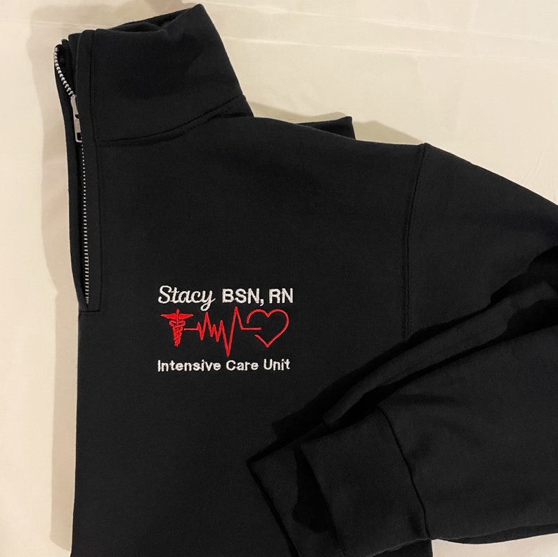 Peut inclure: Veste noire zipp&eacute;e avec le nom "Stacy BSN, RN" brod&eacute; en blanc, ainsi qu'un symbole m&eacute;dical rouge, une ligne de battement de c&oelig;ur et un c&oelig;ur. Le texte "Intensive Care Unit" est en dessous.