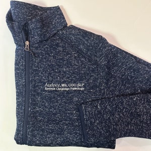 K&ouml;nnte beinhalten: Eine marineblaue und wei&szlig; gesprenkelte Jacke mit Rei&szlig;verschluss und hohem Kragen. Die Jacke hat einen Rei&szlig;verschluss und eine Tasche. Der Text "Audrey, MS, CCC-SLP Speech Language Pathology" ist auf der Vorderseite gestickt.