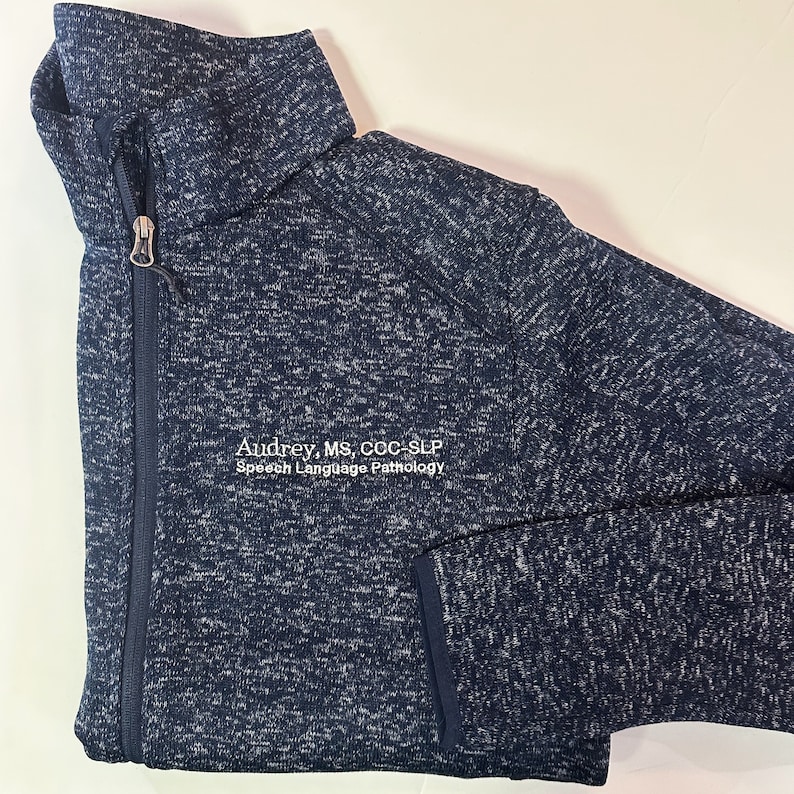 K&ouml;nnte beinhalten: Eine marineblaue und wei&szlig; gesprenkelte Jacke mit Rei&szlig;verschluss und hohem Kragen. Die Jacke hat einen Rei&szlig;verschluss und eine Tasche. Der Text "Audrey, MS, CCC-SLP Speech Language Pathology" ist auf der Vorderseite gestickt.