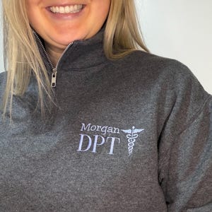 Puede incluir: Una sudadera gris oscuro con cremallera de cuarto de cremallera, con el nombre "Morgan DPT" bordado en blanco, junto con un s&iacute;mbolo de caduceo. La sudadera tiene una cremallera plateada y una textura suave. El dise&ntilde;o est&aacute; en el pecho izquierdo.