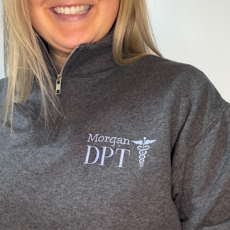 Puede incluir: Una sudadera gris oscuro con cremallera de cuarto de cremallera, con el nombre "Morgan DPT" bordado en blanco, junto con un s&iacute;mbolo de caduceo. La sudadera tiene una cremallera plateada y una textura suave. El dise&ntilde;o est&aacute; en el pecho izquierdo.
