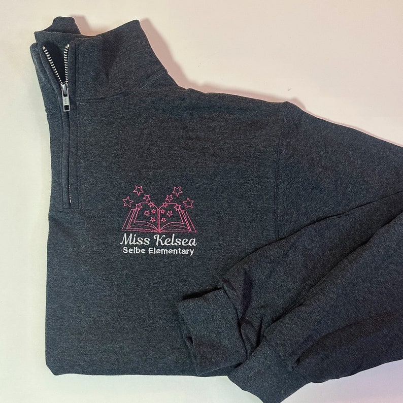 K&ouml;nnte beinhalten: Ein dunkelgrauer 1/4-Rei&szlig;verschluss-Sweatshirt mit einem rosa gestickten Design eines Buches mit Sternen dar&uuml;ber. Der Text "Miss Kelsea Selbe Elementary" befindet sich unter dem Buch.