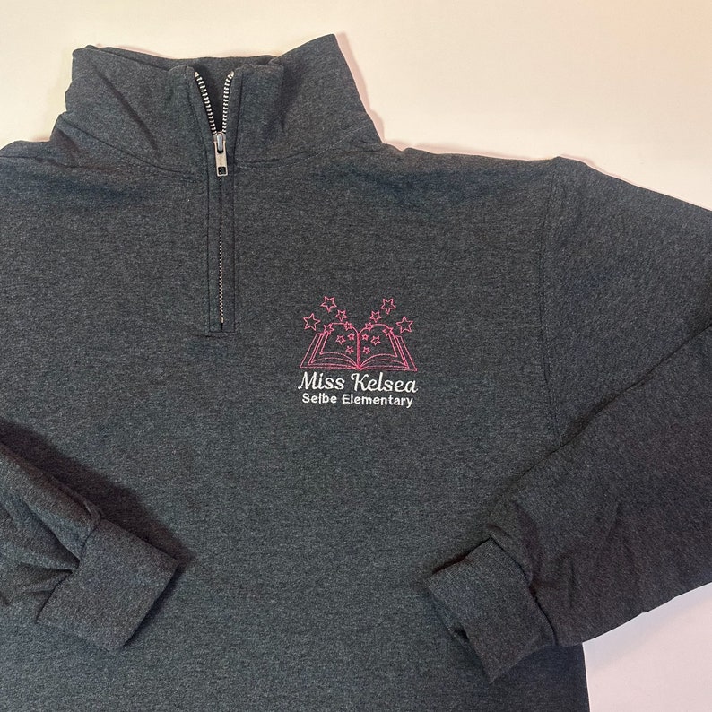 K&ouml;nnte beinhalten: Ein dunkelgrauer 1/4-Rei&szlig;verschluss-Sweatshirt mit einem rosa gestickten Logo, auf dem "Miss Kelsea Seibe Elementary" mit einem Buch- und Sternendesign steht.