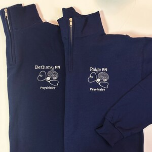 May include: Two navy blue quarter-zip sweatshirts. Each features a white embroidered design of a brain, stethoscope, and the text "Bethany RN Psychiatry" and "Paige RN Psychiatry". The sweatshirts have a ribbed cuff and a gold zipper.