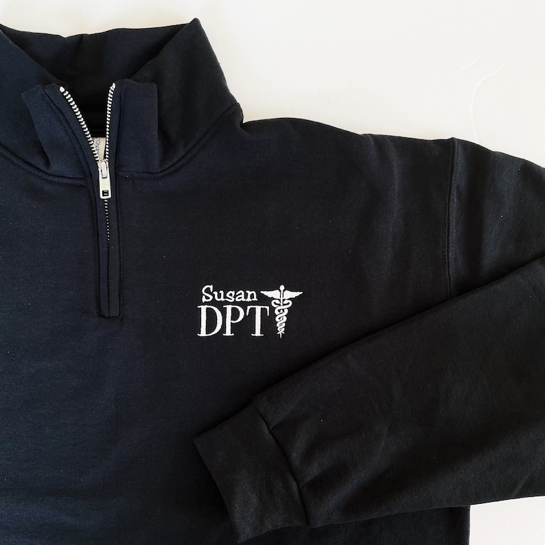 May include: Black quarter-zip sweatshirt with white embroidered text and a medical symbol. The text reads "Susan DPT". The sweatshirt has a high collar and ribbed cuffs. The zipper is silver.