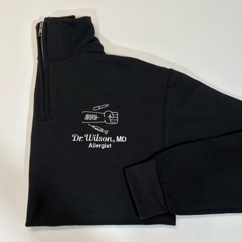 Pode incluir: Camisola preta com fecho de correr de um quarto com texto e gr&aacute;ficos brancos. O texto diz "Dr. Wilson, MD Allergist" abaixo de um gr&aacute;fico de um punho, uma seringa e outras ferramentas m&eacute;dicas.