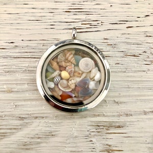 May include: A silver locket necklace with a clear glass insert filled with a variety of small seashells in shades of white, brown, and beige.