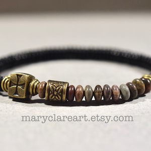May include: A black beaded bracelet with a brass cross charm and decorative beads in shades of brown and gray. The bracelet has a woven appearance and is likely handmade. The Etsy shop name "maryclareart.etsy.com" is visible.