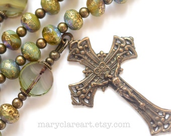 Könnte beinhalten: Nahaufnahme eines Rosenkranzes mit olivgrünen und goldfarbenen Perlen. Der Rosenkranz hat ein detailliertes, bronzefarbenes Kruzifix mit der Figur Jesu Christi. Die Perlen haben eine strukturierte, irisierende Oberfläche.