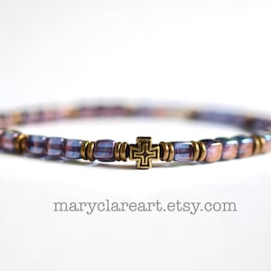 May include: A beaded bracelet featuring iridescent purple square beads alternating with small metallic bronze spacers and a central bronze cross charm. The text maryclareart.etsy.com is printed below the bracelet on a plain white background.