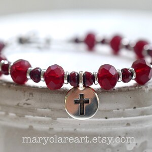 May include: A beaded bracelet featuring faceted, deep red beads and small silver accents. A silver charm with a cross hangs from the bracelet. The website address maryclareart.etsy.com is visible at the bottom.