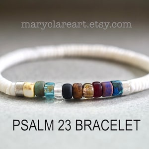 May include: A white bracelet with a series of colourful beads. The beads are in shades of yellow, green, blue, black, brown, red, and purple. The text "PSALM 23 BRACELET" is below the bracelet. The website maryclareart.etsy.com is in the image.