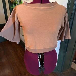 May include: A brown cropped top with a waffle knit texture and short sleeves. The top has a boat neck and is displayed on a mannequin.