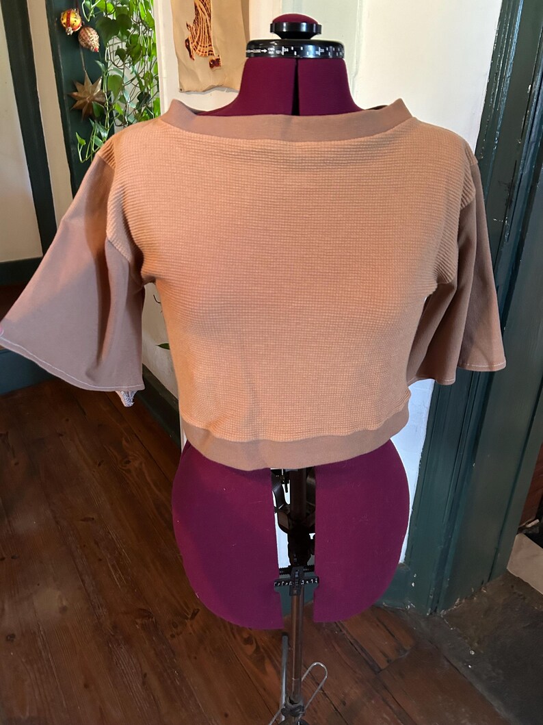 May include: A brown cropped top with a waffle knit texture and short sleeves. The top has a boat neck and is displayed on a mannequin.