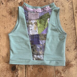 Könnte beinhalten: Ein ärmelloses, mintgrünes Oberteil mit hohem Ausschnitt. Das Mittelteil zeigt ein Patchwork-Design in Lila-, Grün- und Grautönen. Das Oberteil ist aus weichem Stoff gefertigt und hat einen lässigen, künstlerischen Stil.