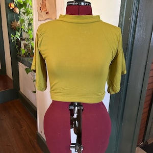 Op de afbeelding: Mosterdgeel crop top met korte mouwen en een col. De top wordt getoond op een rood en zwart paspop. De paspop staat op een metalen standaard. De achtergrond omvat een raam en een muur.