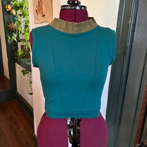 May include: Teal sleeveless crop top with a green ribbed collar. The top is on a dress form.