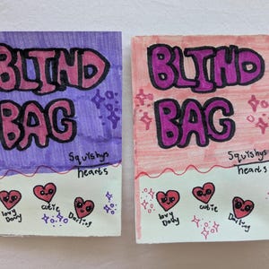 May include: Two handmade "Blind Bag" packages. One is purple, the other is pink. Each has the words "BLIND BAG" in pink bubble letters outlined in black. Below are three red hearts with faces and the words "lovy Dovy", "cutie", and "Darling".