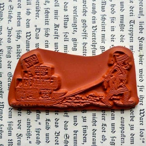 May include: A red rubber stamp with a raised design of a person pulling a cart full of boxes. The person is wearing a long robe and a hat. The cart is on a bumpy road. The stamp is on a white background with text.