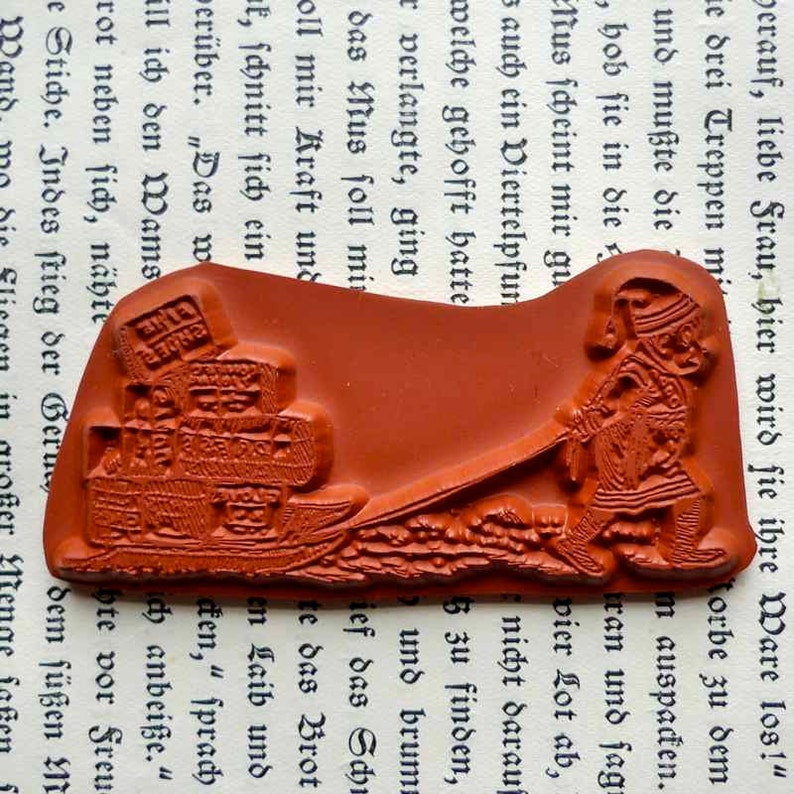 May include: A red rubber stamp with a raised design of a person pulling a cart full of boxes. The person is wearing a long robe and a hat. The cart is on a bumpy road. The stamp is on a white background with text.