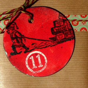 May include: Red circular Christmas ornament with a black and white illustration of a person pulling a cart full of presents. The ornament has a white circle with the number 11 in the center.