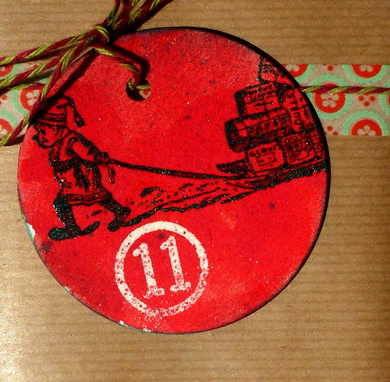 May include: Red circular Christmas ornament with a black and white illustration of a person pulling a cart full of presents. The ornament has a white circle with the number 11 in the center.