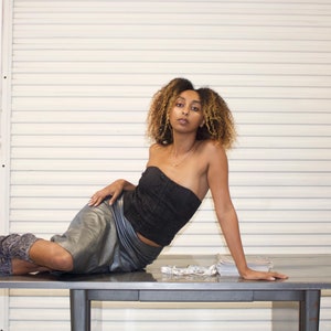 May include: A person wearing a black strapless top, silver skirt, and patterned knee-high boots, reclining on a metal table. The background features a white shuttered wall and a window with grey curtains.