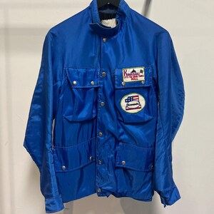 May include: A bright blue jacket with multiple front pockets and snap closures. The jacket features two patches: one with text and a mountain graphic, and another with an American flag design. The jacket is hanging on a metal rack.
