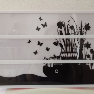 Könnte beinhalten: Ein weißes beleuchtetes Schild mit einer schwarzen Silhouette von Blumen und Schmetterlingen. Die Blumen stehen in einer Vase und die Schmetterlinge fliegen um sie herum.