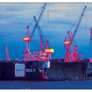 Könnte beinhalten: Ein großes, schwarzes Schiff liegt in einem Schiffbauunternehmen an. Das Schiff ist von mehreren großen, rosafarbenen Kränen mit gelben Akzenten umgeben. Das Schiff trägt die Aufschrift "Blohm+Voss" und "DOCK 11" auf der Seite.